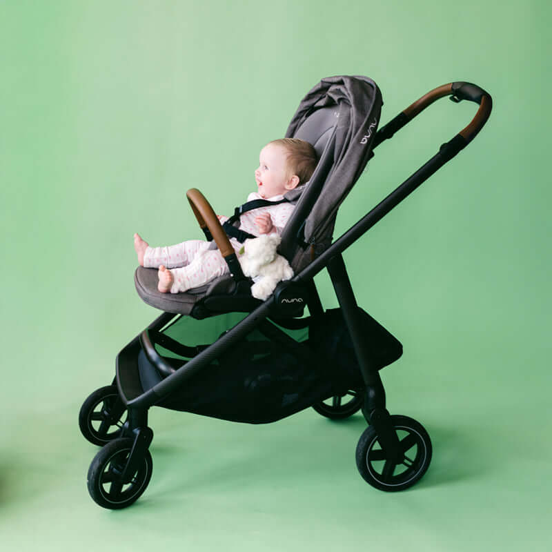 A baby in a gray and black stroller with brown handle accents, photographed against a solid light green studio background.