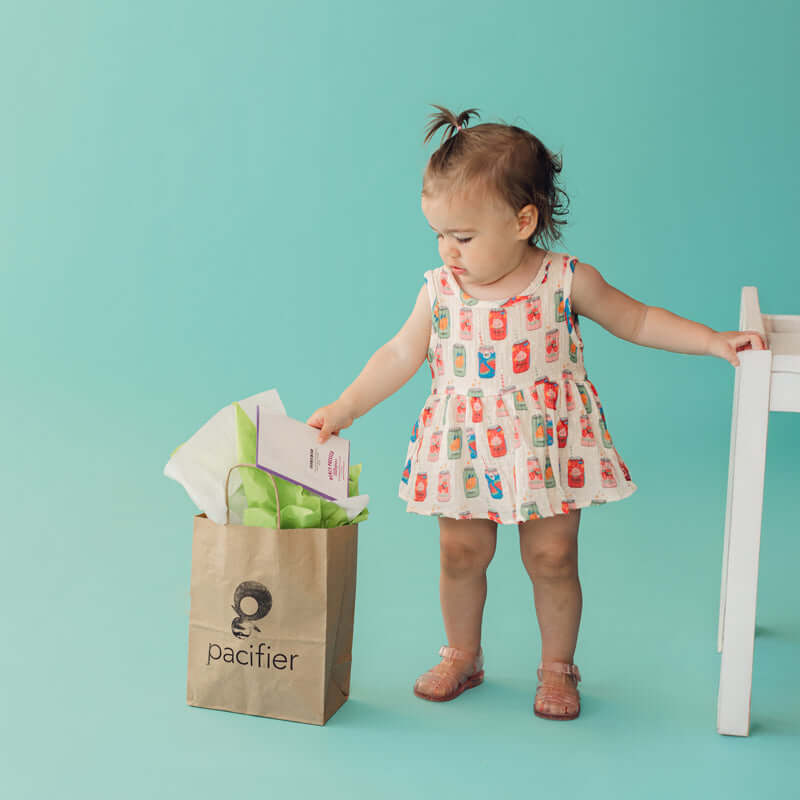 A toddler girl stands next to a brown paper bag printed with the 'pacifier' logo, holding tissue paper inside. She is wearing a sleeveless white dress with a colorful milk carton pattern and is posed against a solid turquoise background.