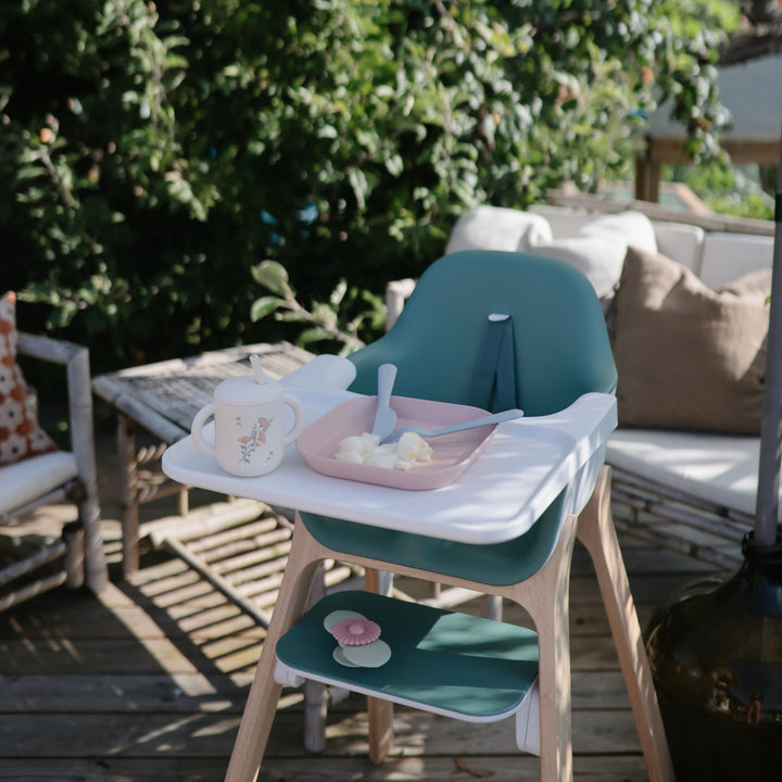 High chair with a tray containing a cup, plate, and utensils on a wooden deck with greenery in the background.