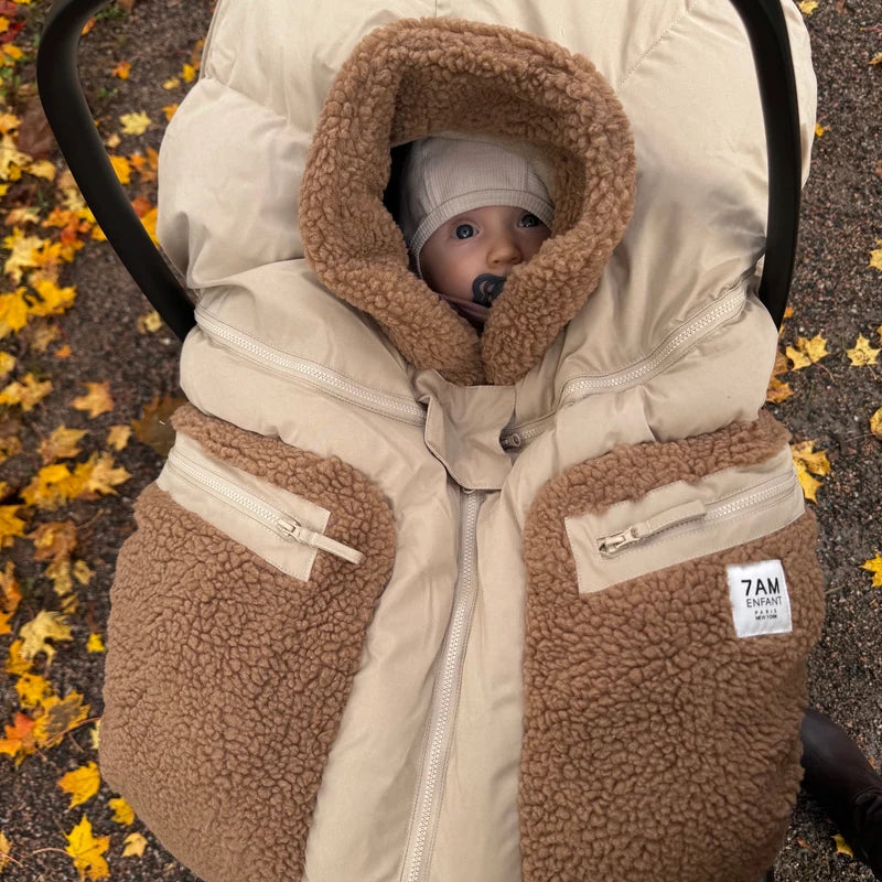 A baby wearing a beanie and pacifier is cozy inside an infant car seat outfitted with the Benji Applique Car Seat Cocoon by 7AM Enfant in oat with brown sherpa fleece trim, set against a backdrop of autumn leaves on the ground.