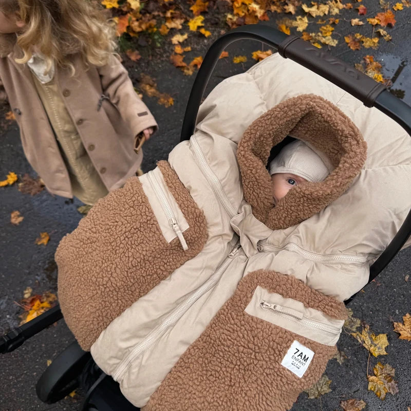 A young child with a plaid hair bow reaches down toward an infant car seat where a baby is sleeping inside a Benji Applique Car Seat Cocoon by 7AM Enfant in oat with brown sherpa fleece trim, outdoors near a paved path scattered with autumn leaves.