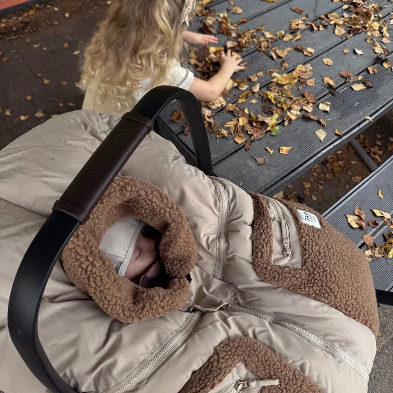 A young child with long blonde hair reaches down toward an infant car seat where a baby is sleeping inside a Benji Applique Car Seat Cocoon by 7AM Enfant in oat with brown sherpa fleece trim, outdoors near a wooden bench scattered with autumn leaves.