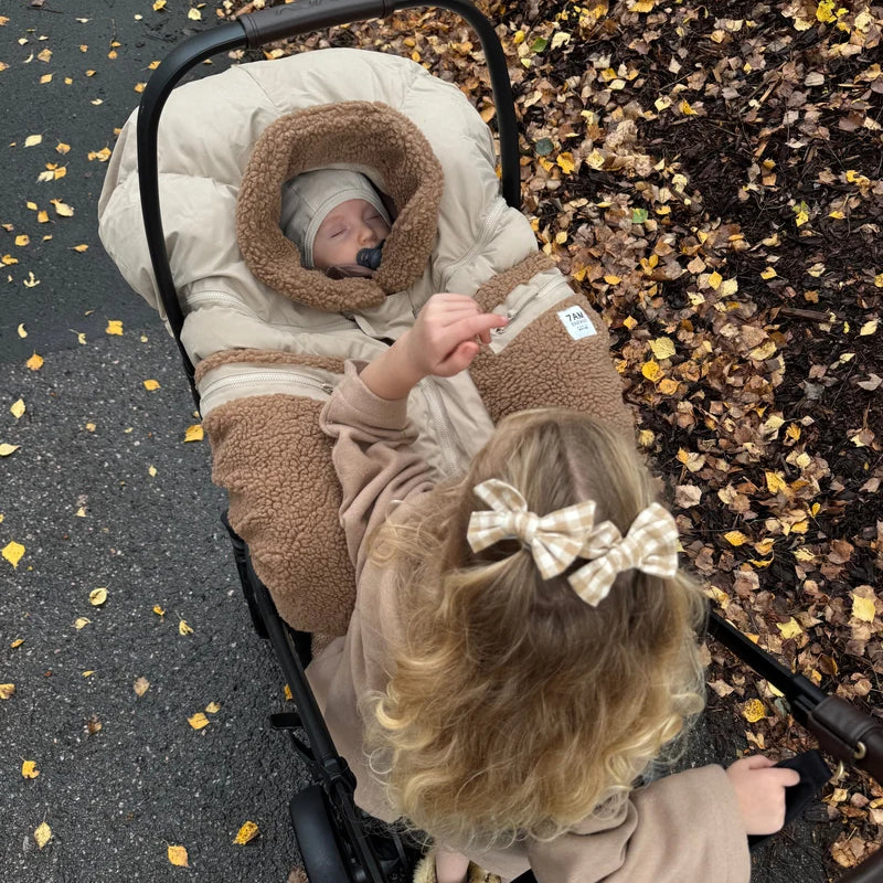 A young child with a plaid hair bow reaches down toward an infant car seat where a baby is sleeping inside a Benji Applique Car Seat Cocoon by 7AM Enfant in oat with brown sherpa fleece trim, outdoors near a paved path scattered with autumn leaves.