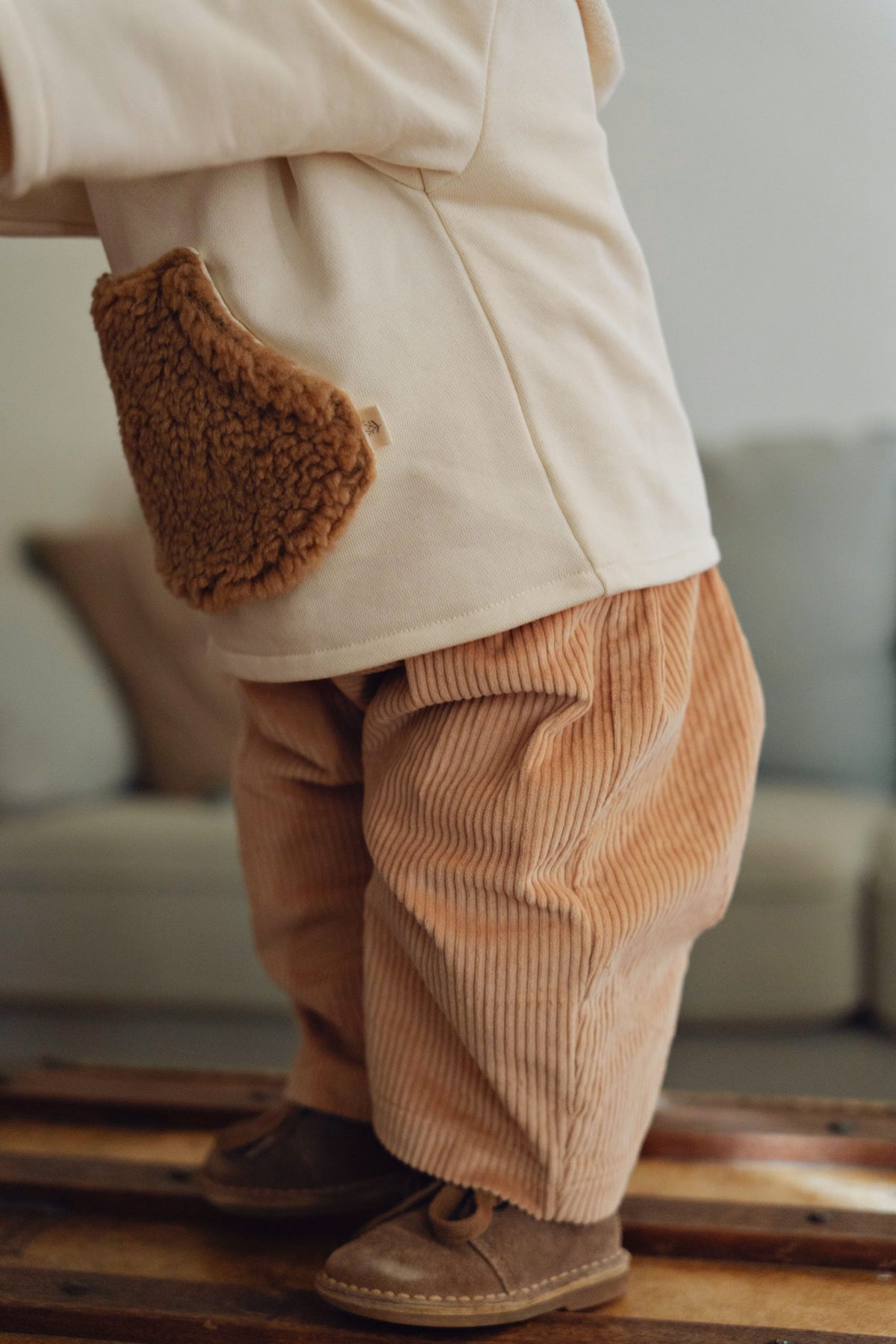 A close-up lifestyle shot of a child wearing beige corduroy pants with a brown sherpa-lined patch pocket on the back, standing on a wooden floor.