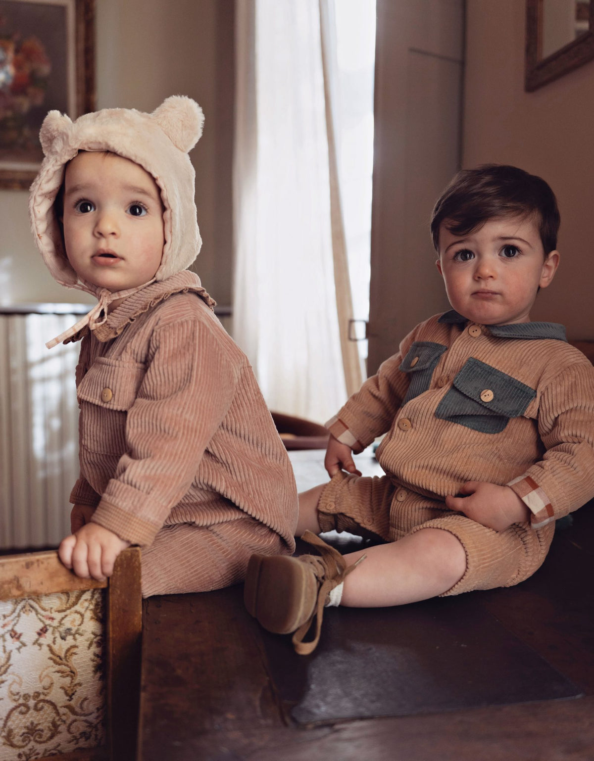 Two toddlers wearing matching corduroy outfits, seated indoors on a wooden bench. The child on the left wears a beige hooded romper with bear ears, and the child on the right wears a matching corduroy romper with dark green chest pockets.