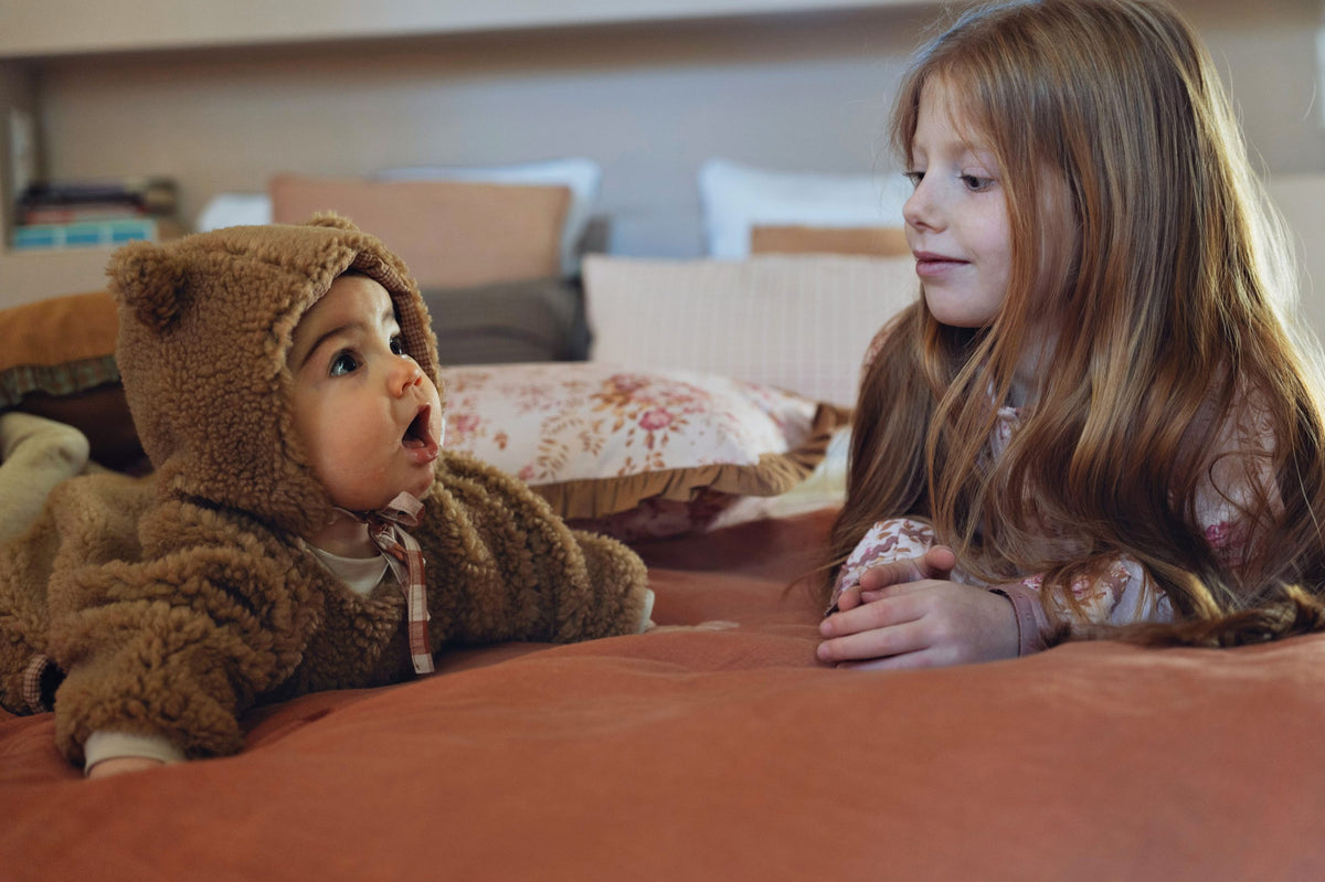A baby and a girl lying on a rust-colored fabric surface wearing a brown teddy fleece hooded outfit with ear details and drawstrings. A matching brown fleece hood is also shown nearby.background.