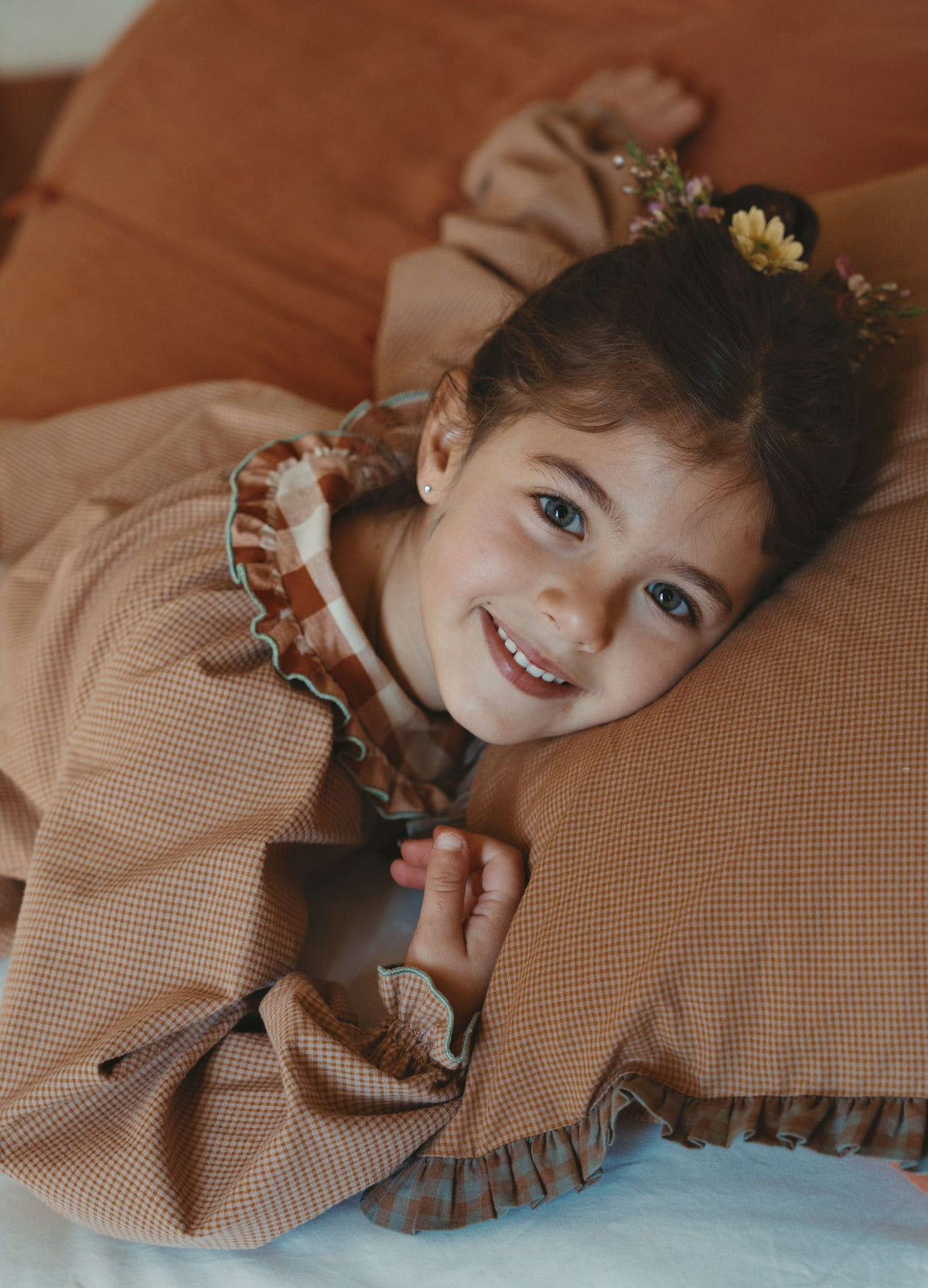 A young girl with dark hair and a small flower hair accessory lies on a brown checkered pillow, wearing a matching brown checkered long-sleeve top with a ruffled collar.