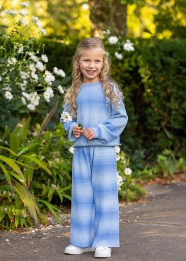 A young girl with blonde hair stands outdoors on a path wearing a matching light blue two-piece lounge set consisting of a long-sleeve top and wide-leg pants, with white sneakers.