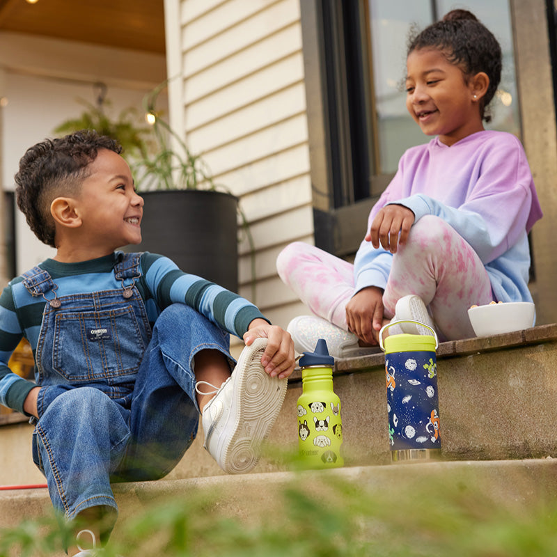 Two children sitting on a step outdoors with colorful water bottles.