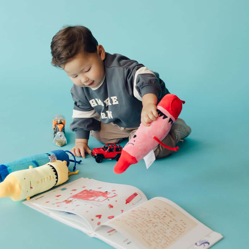 A young child sits on a light blue floor playing with a pink plush toy, an open book, and small toys against a solid light blue background.