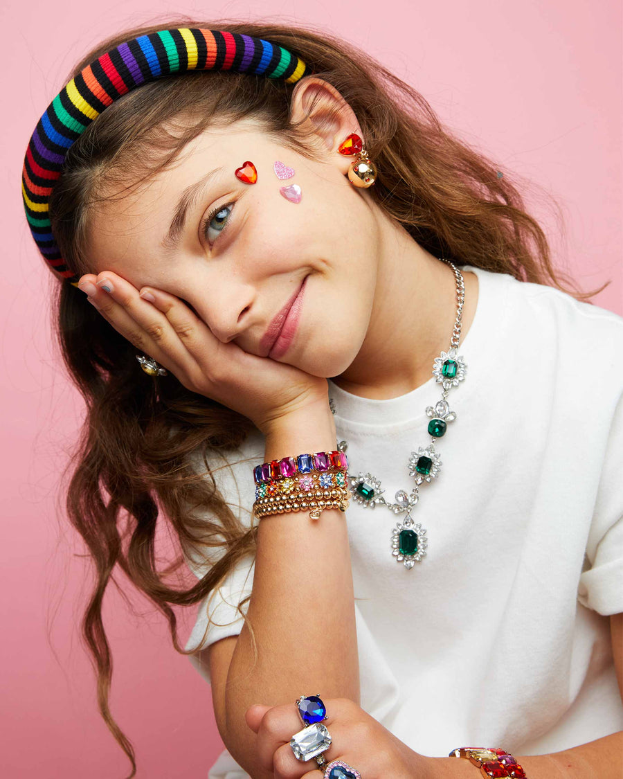 Young girl with colorful jewelry including a rainbow headband, necklaces, bracelets, and rings on a pink background.