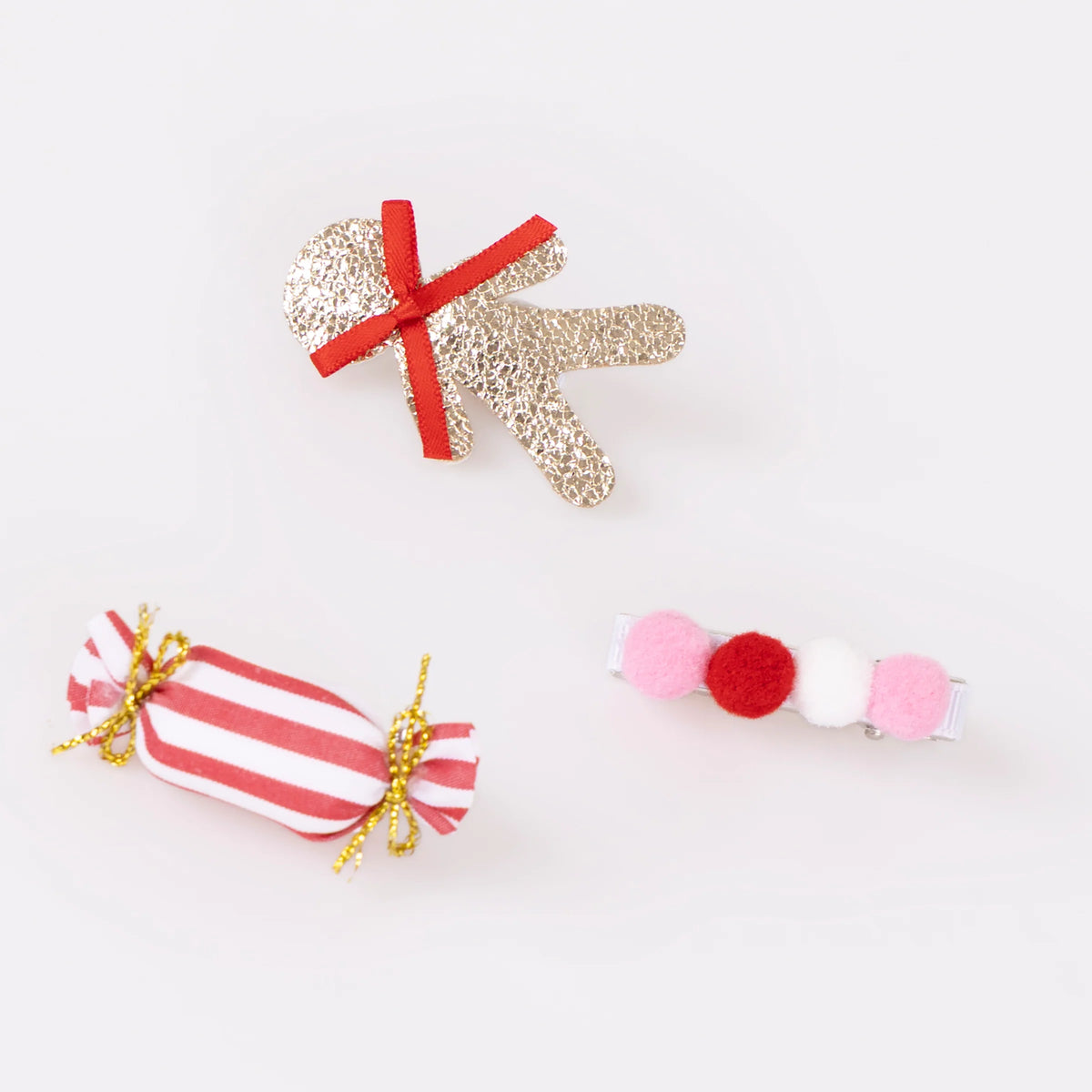 A set of festive holiday-themed hair clips featuring a sparkly gingerbread person, a striped candy, and colorful pompoms, resembling accessories by Meri Meri.