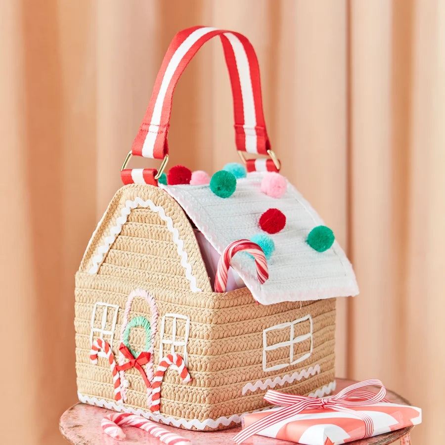 A straw house-shaped handbag with a red and white striped handle, decorated with candy canes, colorful pom-poms, and white lace-like trim, set against a soft peach curtain background.