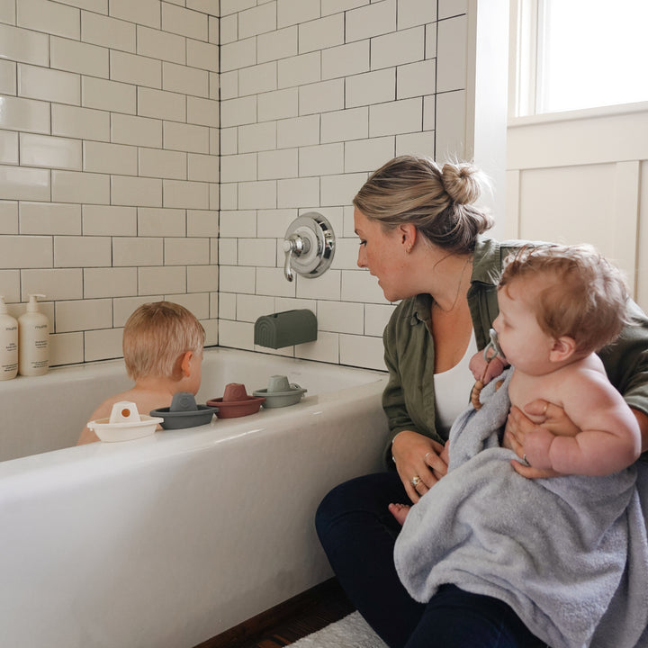 Woman holding a baby while another child sits in a bathtub with toys.