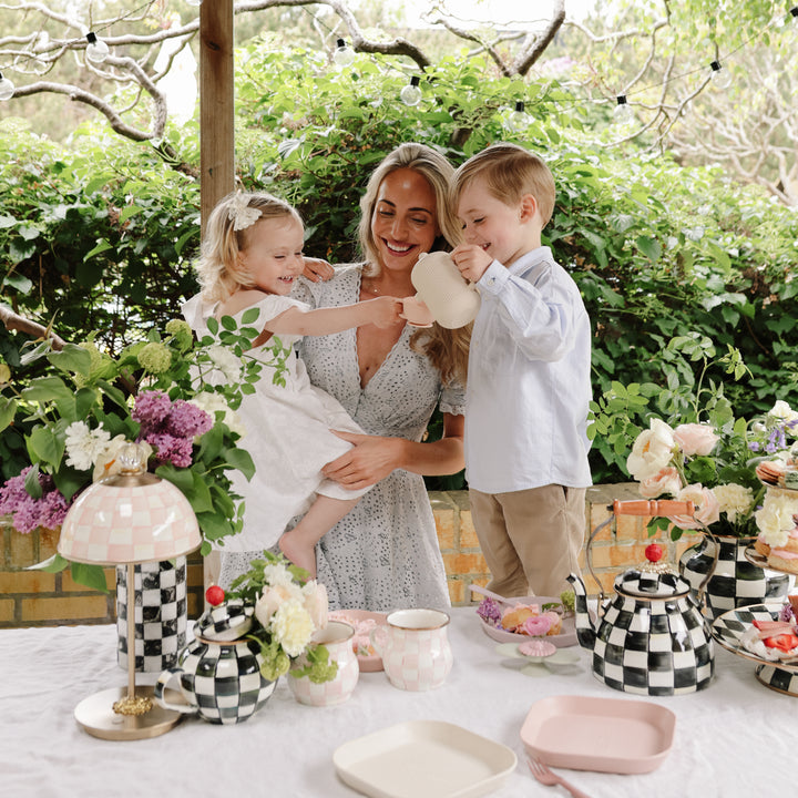 A woman holding a young child outdoors next to an outdoor table set up with flowers, tableware, and a toy tea set, while a boy stands nearby holding a toy teapot.