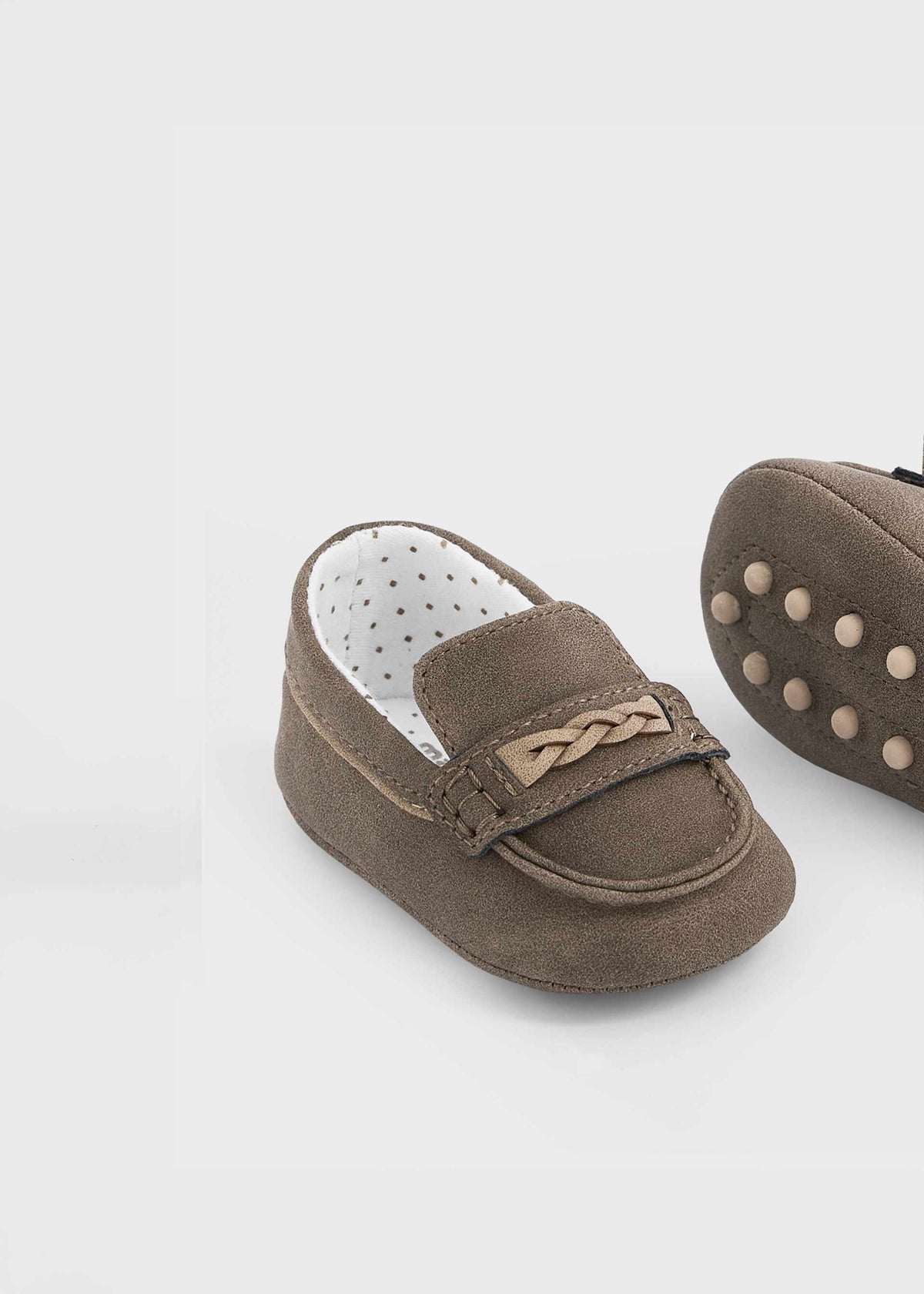 A pair of baby loafers in taupe brown suede, featuring a decorative braided strap on the vamp and a white dotted interior lining. One shoe is shown from the side profile, and the other is partially visible from the sole showing circular grip dots, set against a clean white background.