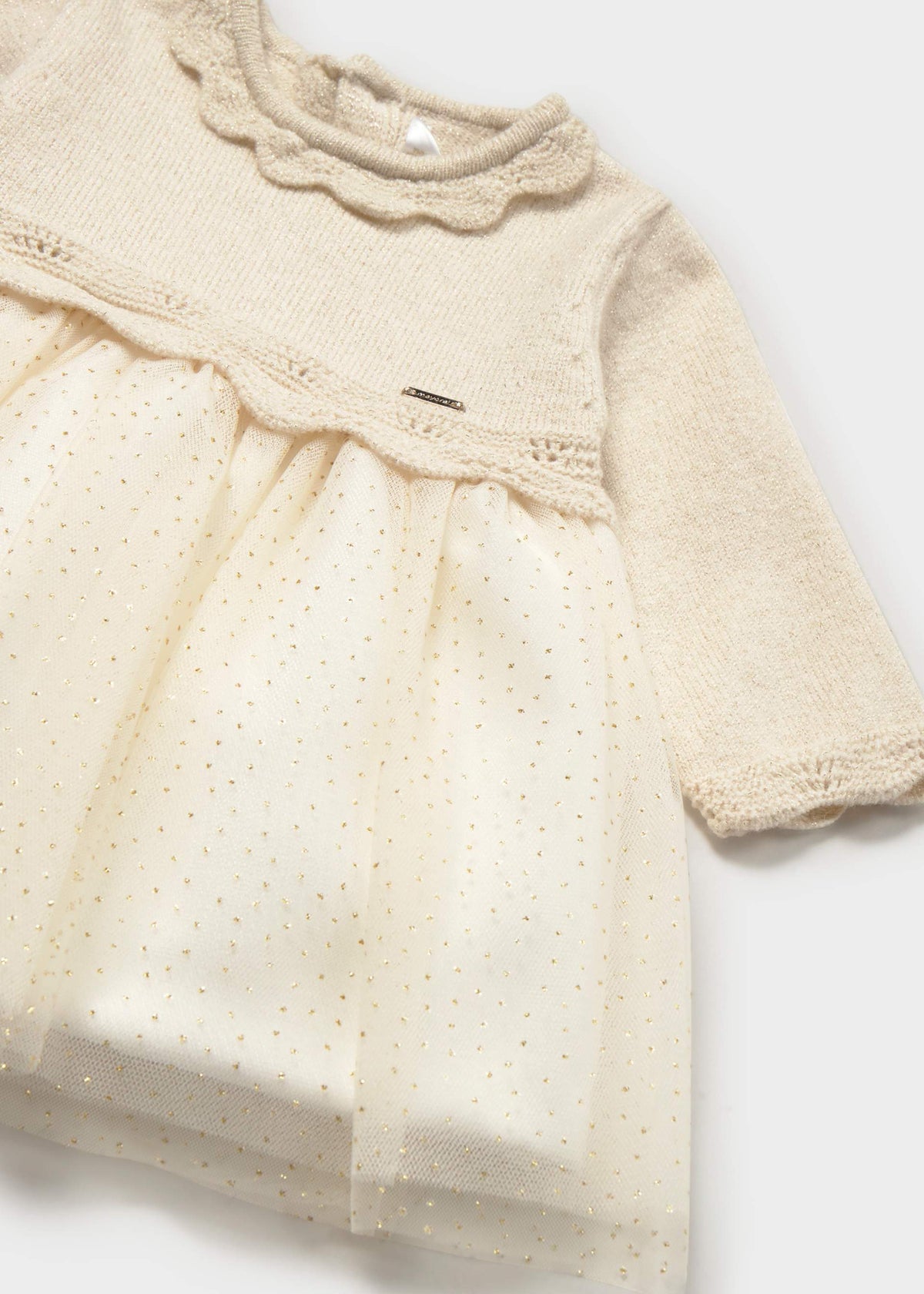 A close-up of a baby dress in cream with a textured knit bodice, scalloped collar, long sleeves, and a sheer eyelet tulle skirt with tiny gold polka dots.