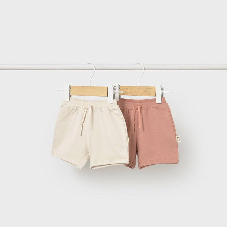 Two pairs of shorts, one beige and one pink, hanging on a white background.