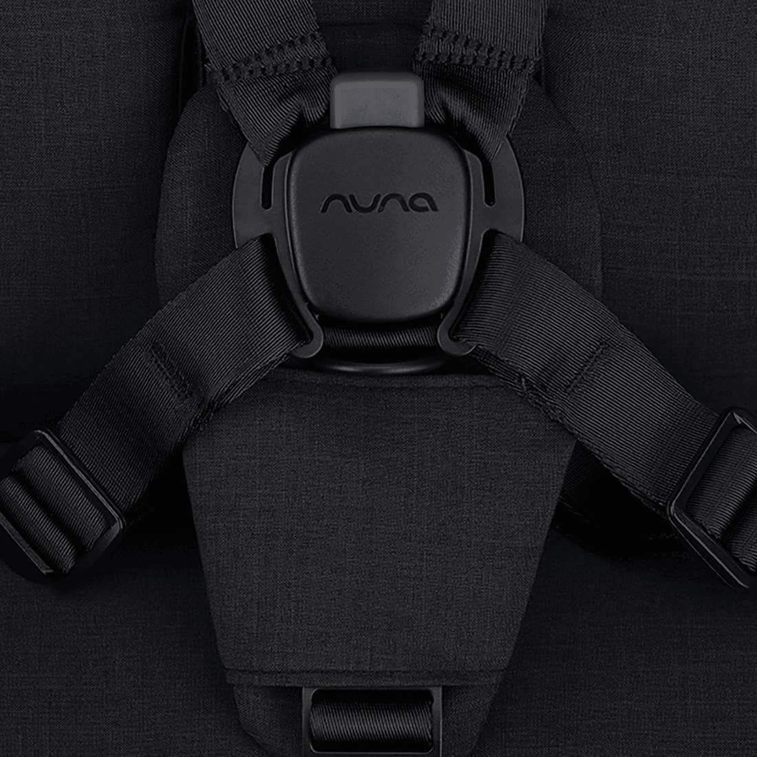 Close-up of a black car seat with Nuna logo on the buckle area against a dark background.
