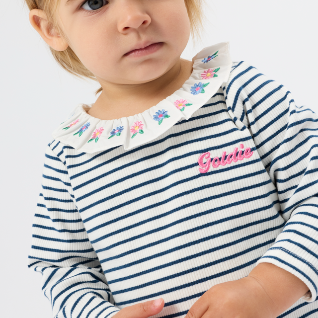 A close-up of a young child wearing a navy and white horizontally striped long-sleeve top with a white embroidered floral collar and a pink 'Gabbie' script on the chest, against a light gray background.