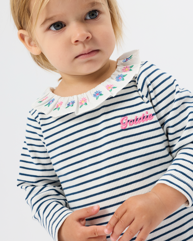 A close-up of a young child wearing a navy and white horizontally striped long-sleeve top with a white embroidered floral collar and a pink 'Gabbie' script on the chest, against a light gray background.