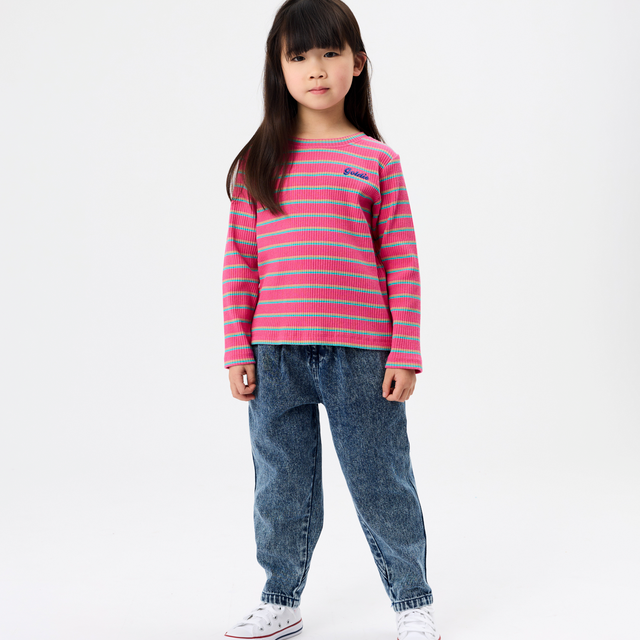 A young girl stands against a plain white studio background wearing a pink and light blue horizontally striped long-sleeve knit top, blue jeans, and white sneakers.
