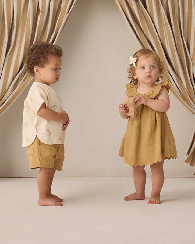 Two children in matching yellow outfits standing in front of a beige curtain.
