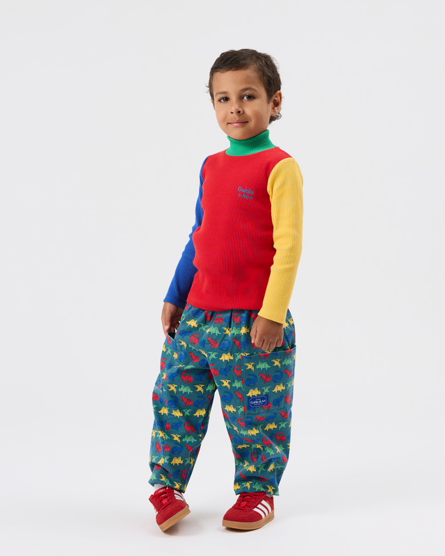 A young child modeling a color-blocked knit sweater vest with long yellow and blue sleeves over a green mock neck, paired with colorful dinosaur-print pants and red sneakers, on a light gray studio background.