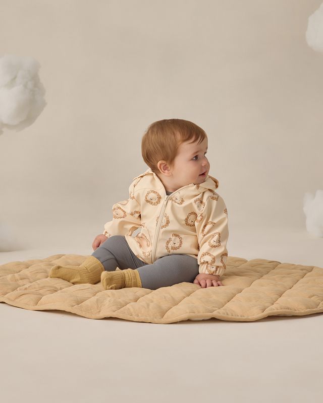 Baby in a beige quilted jacket sitting on a beige blanket against a neutral background