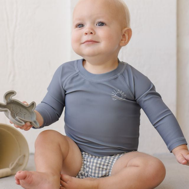 Baby wearing a blue long-sleeve shirt and checkered diaper sitting on a light-colored surface.