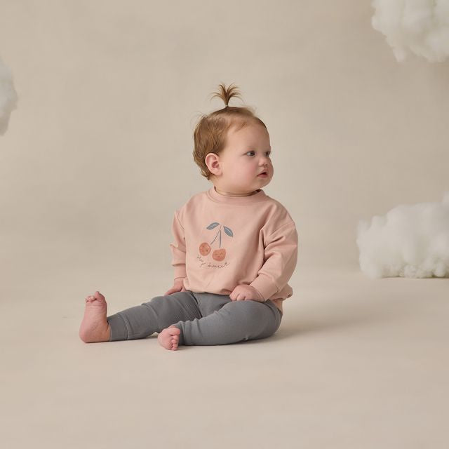 Baby sitting on a beige surface wearing a pink sweatshirt with a cherry design and gray pants.