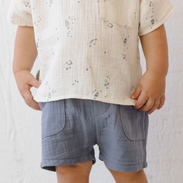 Child wearing a beige shirt with a subtle pattern and blue shorts on a plain background