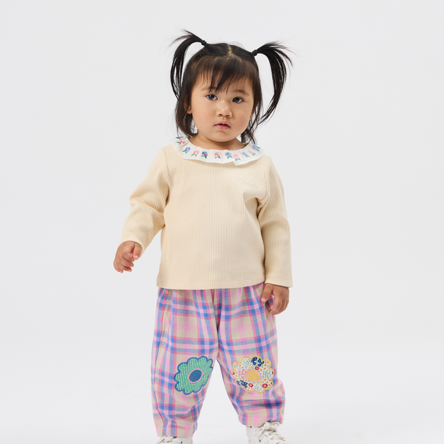 A toddler girl with black pigtails stands against a light gray studio background wearing a cream long-sleeve top with a blue patterned collar, pink and purple plaid pants with embroidered flowers, and white sneakers.