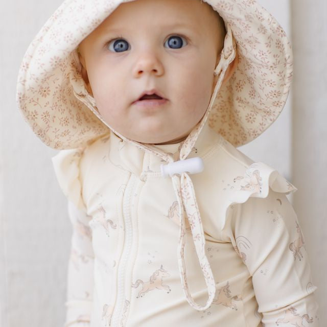 Baby wearing a cream-colored outfit with a matching hat against a light background