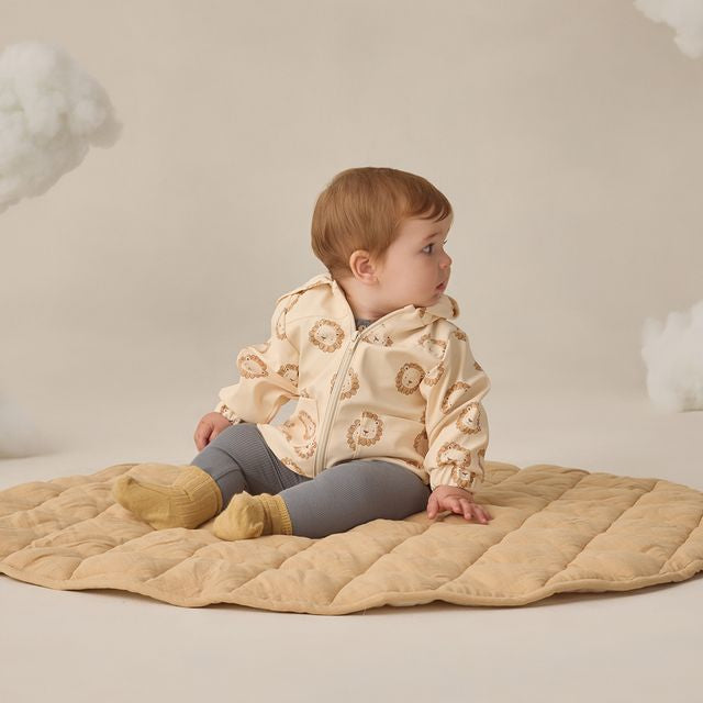 Baby sitting on a beige quilted blanket wearing a cream jacket with floral patterns against a neutral background.