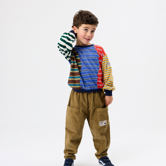 A young boy modeling a colorful patchwork sweatshirt with green, blue, red, and yellow striped panels, paired with brown corduroy pants and navy sneakers, against a plain white studio background.