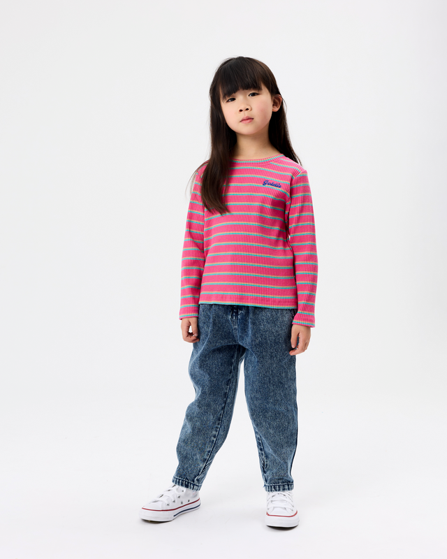 A young girl stands against a plain white studio background wearing a pink and blue horizontally striped long-sleeve knit top, blue jeans, and white sneakers.