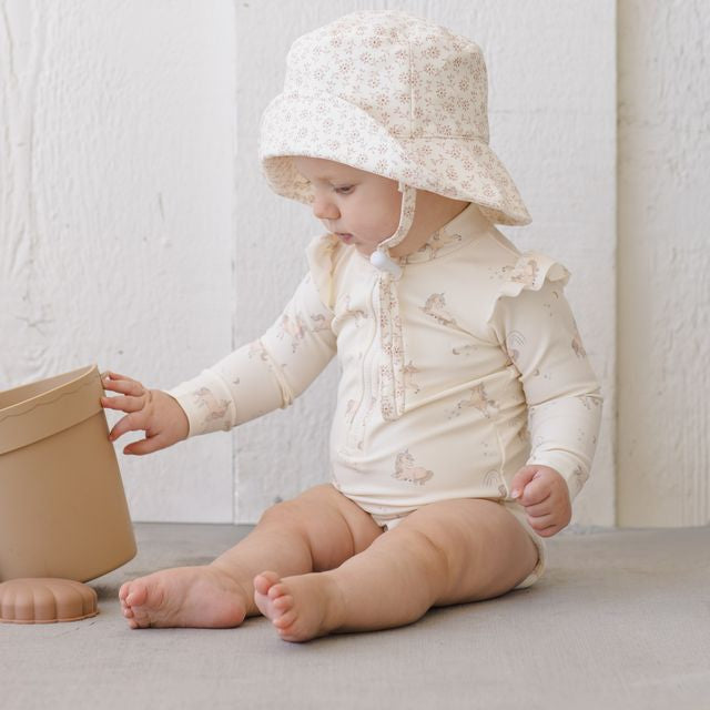 Baby in a white outfit and floral hat playing with a beige pot on a light surface.