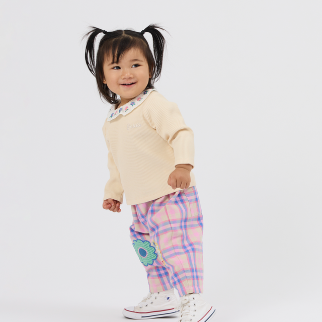 A toddler girl wearing a cream long-sleeve top, pink and purple plaid pants with a blue flower applique, and white canvas sneakers, standing against a light gray studio background.