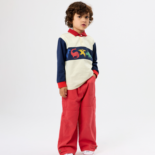 A child model wearing a color-blocked long-sleeve polo shirt with a colorful graphic across the chest and matching red wide-leg trousers, standing against a plain white studio background.