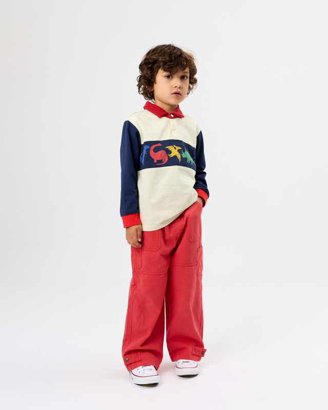 A child model wearing a color-blocked long-sleeve polo shirt with a colorful graphic across the chest and matching red wide-leg trousers, standing against a plain white studio background.