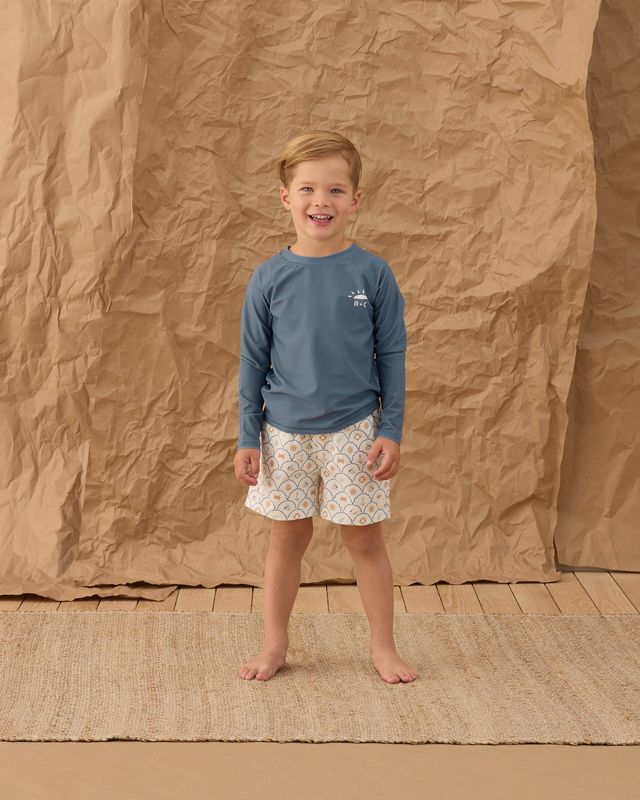 Child wearing a blue long-sleeve shirt and patterned shorts standing against a brown textured backdrop.