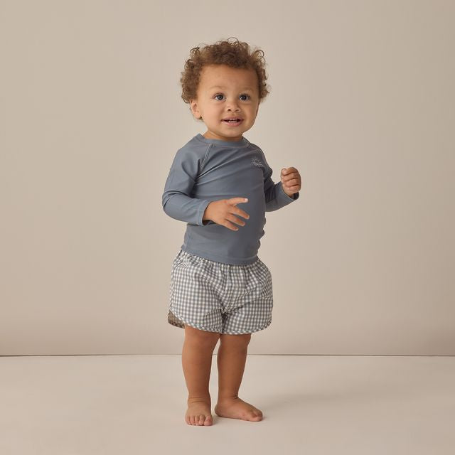 Child wearing a blue long-sleeve shirt and checkered shorts on a beige background