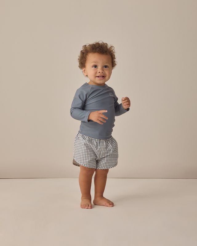 Child wearing a blue long-sleeve shirt and checkered shorts on a beige background