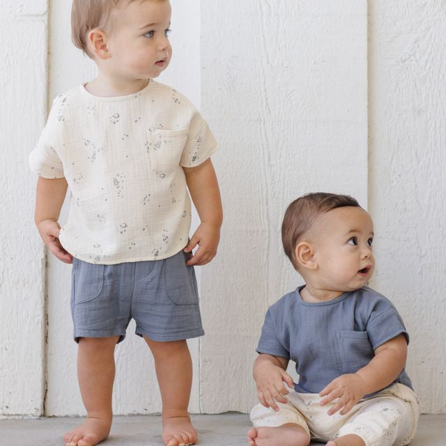Two toddlers standing against a white wall, one wearing a white shirt with small patterns and blue shorts, the other in a blue shirt and beige pants.