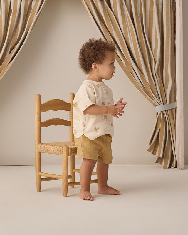 Child standing next to a wooden chair in a room with striped curtains.