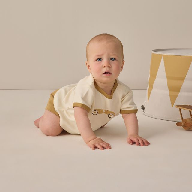 Baby in a white and brown onesie sitting on a light-colored floor with a neutral background
