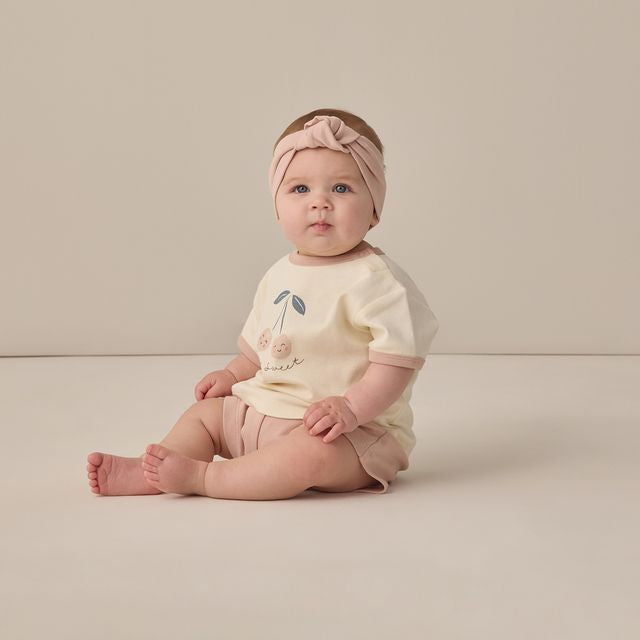 Baby sitting on a plain background wearing a light-colored outfit with a headband.