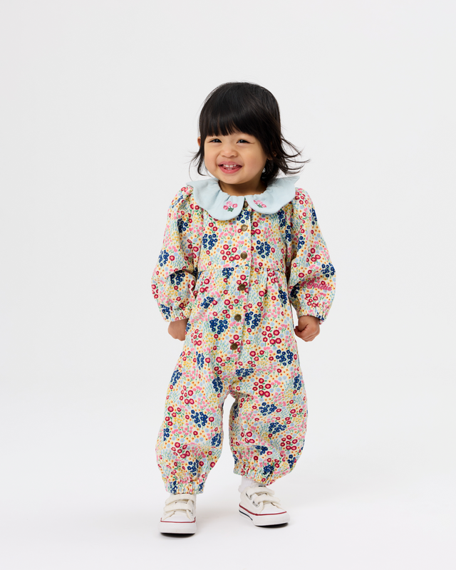 A toddler girl wearing a cream-colored floral jumpsuit with a light blue Peter Pan collar, standing against a plain white studio background.