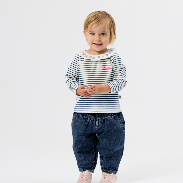 A toddler girl standing against a white studio background, wearing a navy and white striped long-sleeve top with a ruffled collar, dark blue denim pants, and light pink sneakers.
